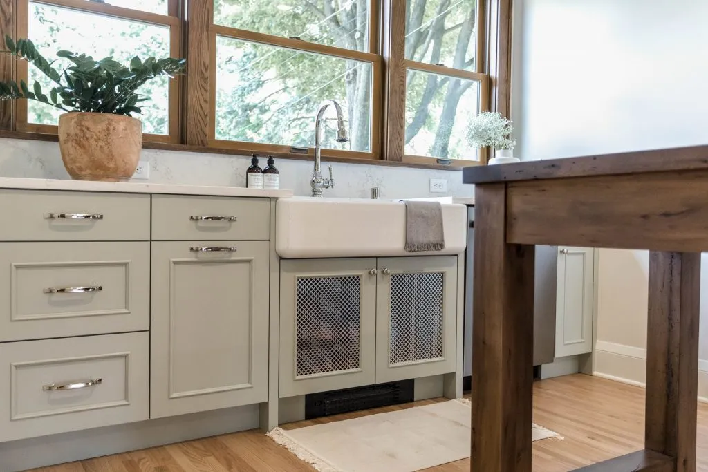 Custom green kitchen cabinetry in Tudor style home with white apron front sink, polished nickel hardware, and wood windows