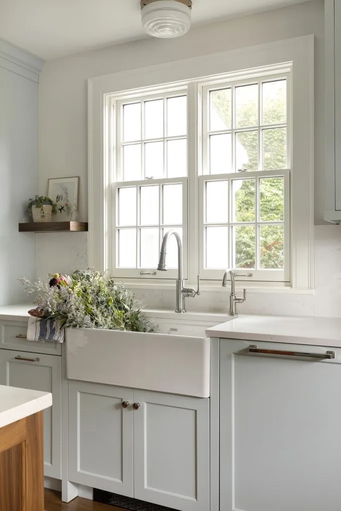 Custom light blue kitchen cabinetry with large apron front farmhouse sink in Colonial kitchen with vintage light fixtures, paneled dishwasher, polished nickel cabinet hardware