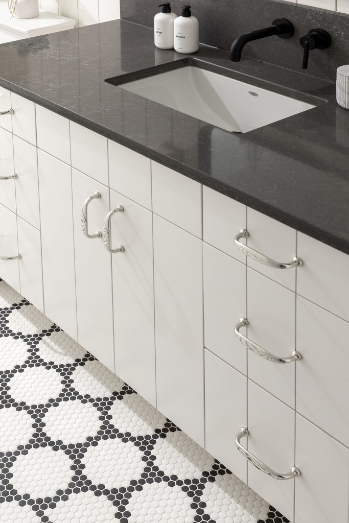 Custom white painted bathroom vanity with kerf cabinet doors, polished nickel hardware, black wall mount faucet, dark grey quartz countertop, glass penny mosaic floor tile