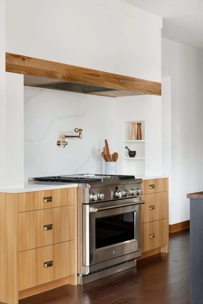 Custom red elm cabinetry with white alcove range hood and reclaimed wood band, quartz backsplash and brass pot filler