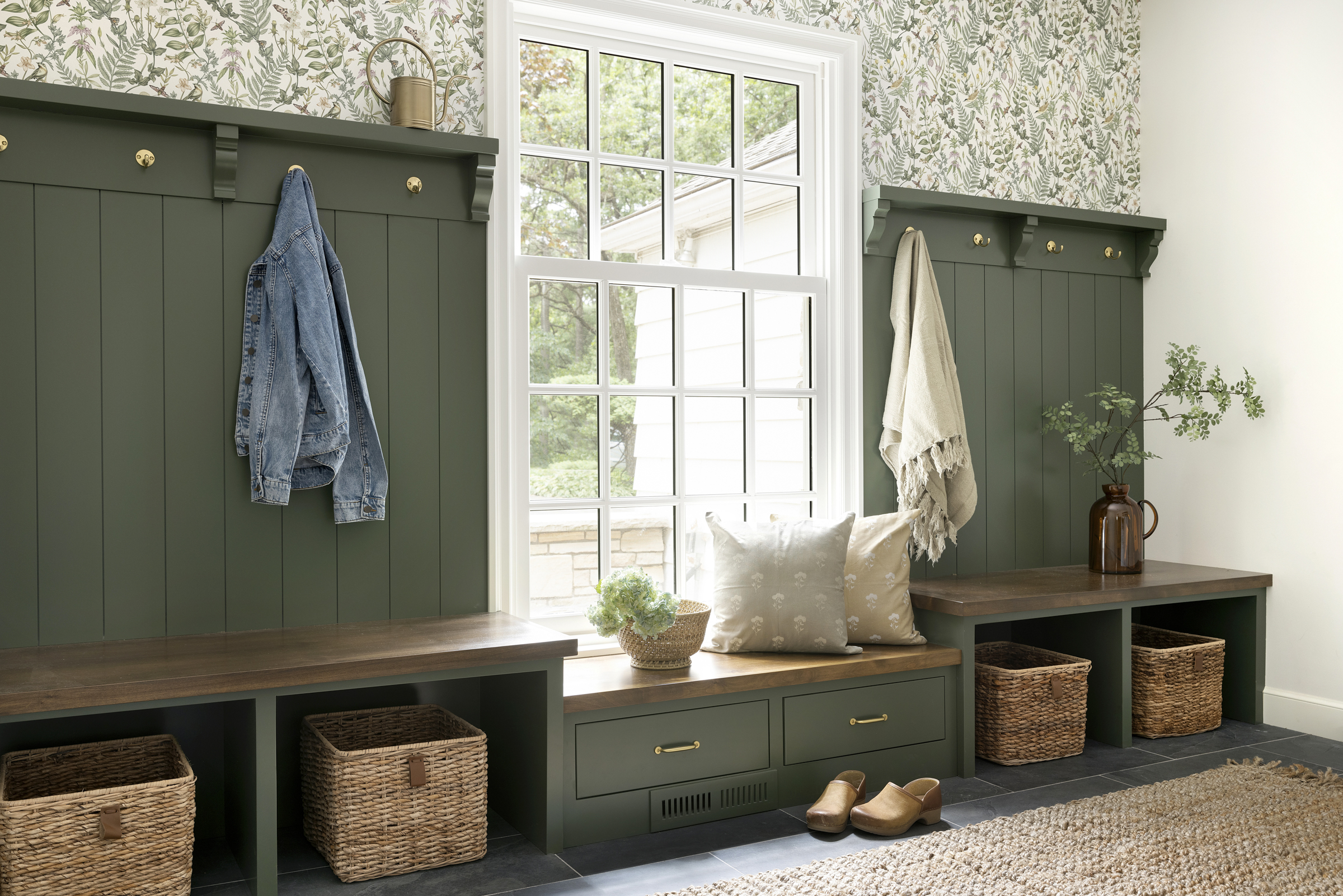 Open mudroom storage with brass hooks, and wallpaper paired with green cabinetry.