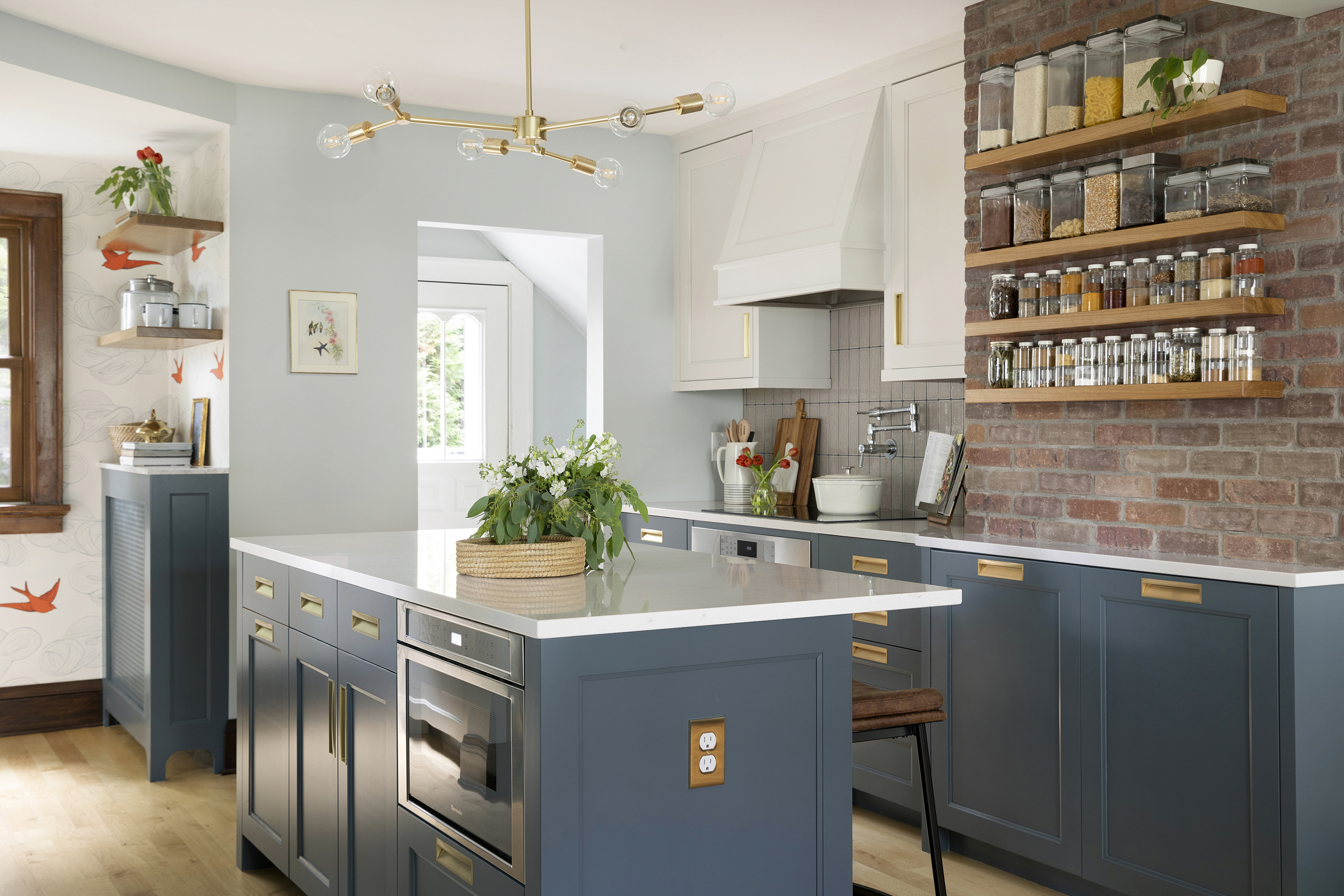 Blue and white custom kitchen with brass recessed cabinet hardware and lighting, orange bird wallpaper, island in small kitchen, brick chimney wall with wood shelves for spice display