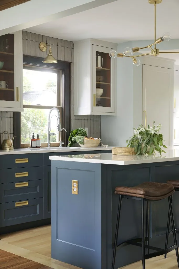 Blue and white custom kitchen cabinets with orange behind glass cabinet doors, industrial style sink faucet, brass window sconce and vertical taupe backsplash tile