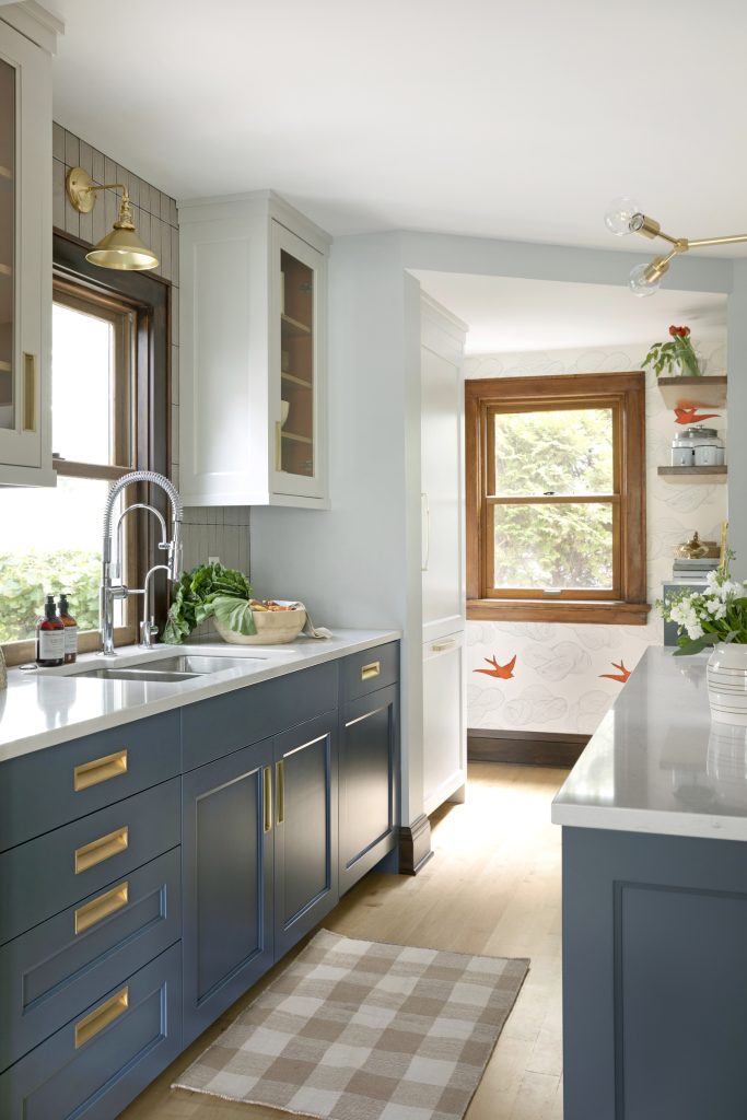 Blue and white custom kitchen cabinets with orange behind glass cabinet doors, industrial style sink faucet, brass window sconce and vertical taupe backsplash tile