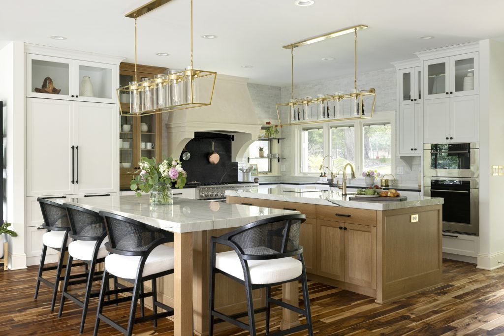 Custom kitchen renovation featuring double white oak islands with overhead brass linear chandeliers and natural quartzite countertops, white perimeter custom cabinetry with natural soapstone countertops, black cabinet hardware and custom plaster range hood.