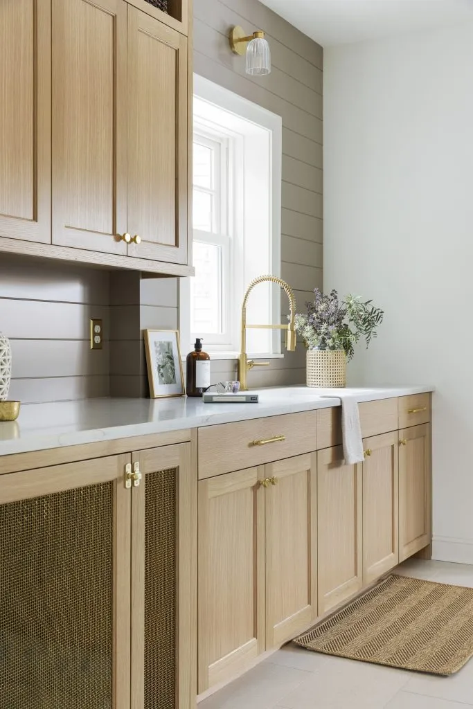 A modern mudroom drop zone features white oak wood cabinets with gold hardware, a light countertop, a brass faucet, a window, with a painted shiplap paneled wall.