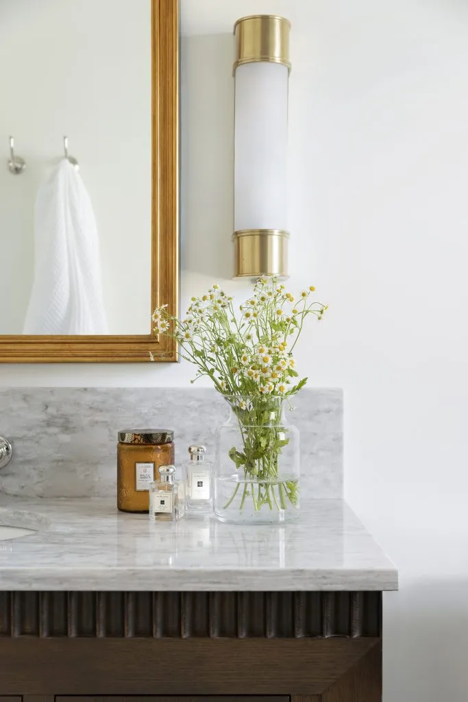 A bathroom countertop with a marble surface showcases the elegance of natural stone. Small perfume bottles, a brown candle, and fresh daisies in a glass vase complement the scene. Above, a wooden-framed mirror and brass wall sconce complete the look.