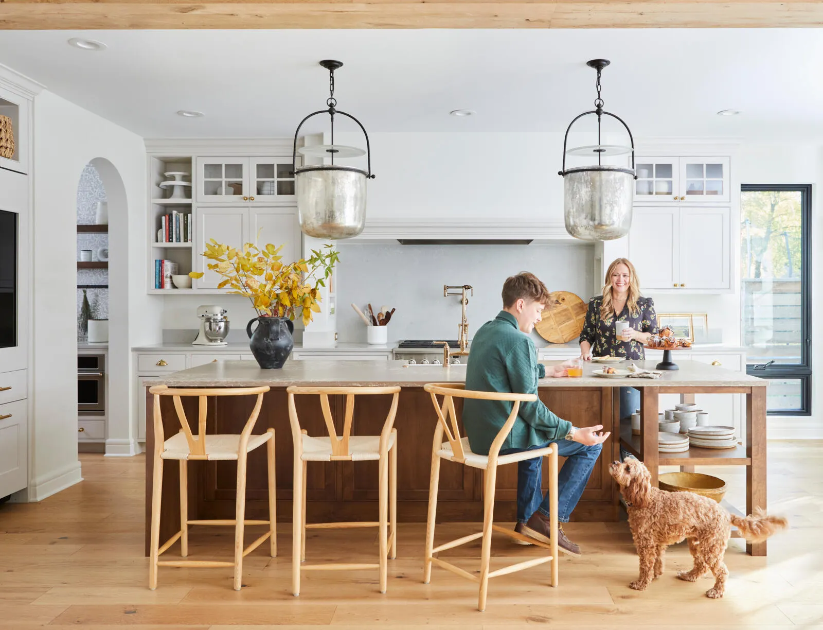 A bright, modern kitchen with Why Choose Natural Stone Countertops featured at the island, where a man sits as a woman prepares food and a small brown dog looks up. The space boasts wood floors, light cabinets, and large pendant lights.