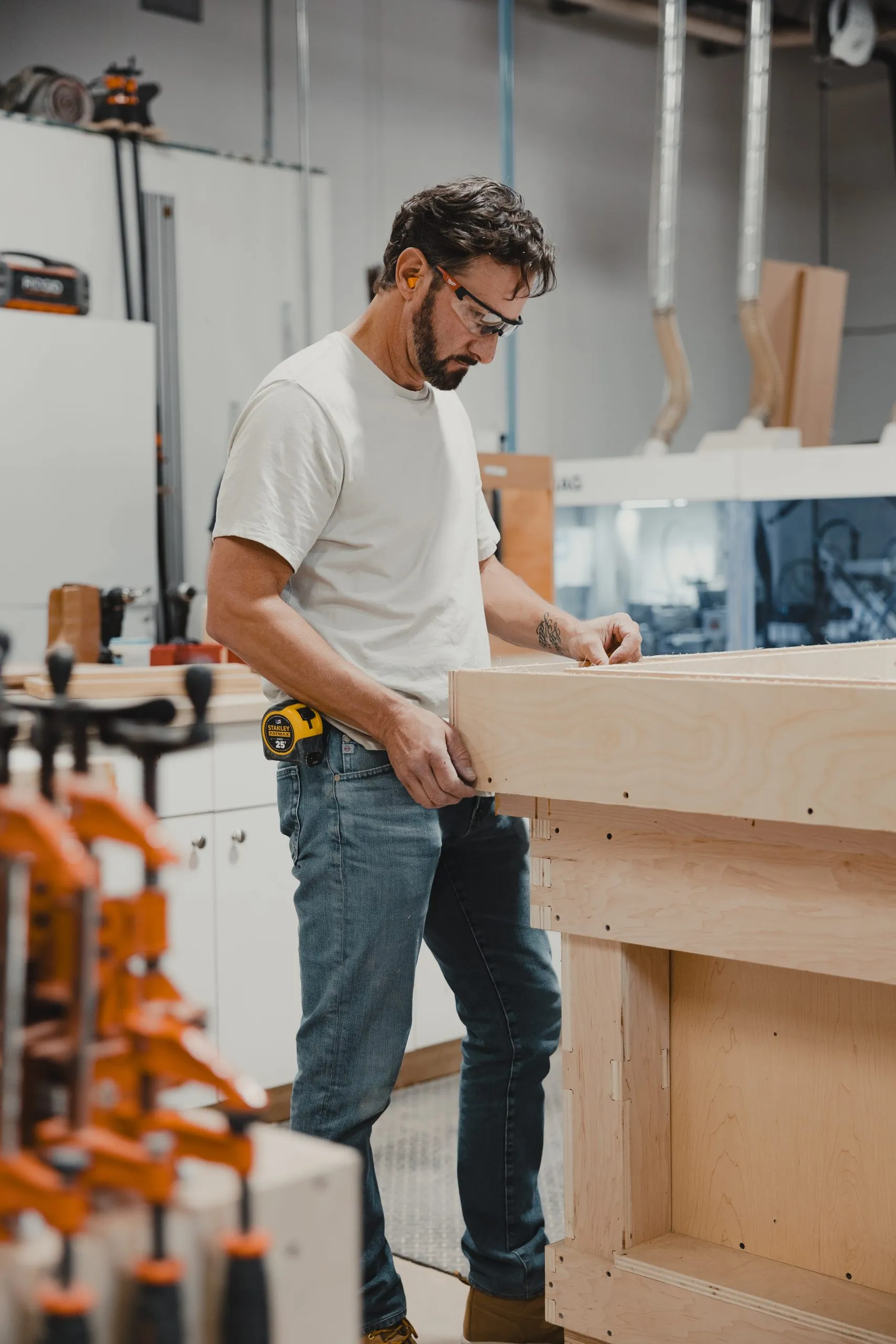 Jesse wearing safety glasses and earplugs works with a large wooden structure in a workshop, surrounded by tools and clamps—crafting his project with focus, perhaps creating one of the perfect Fathers Day gifts.