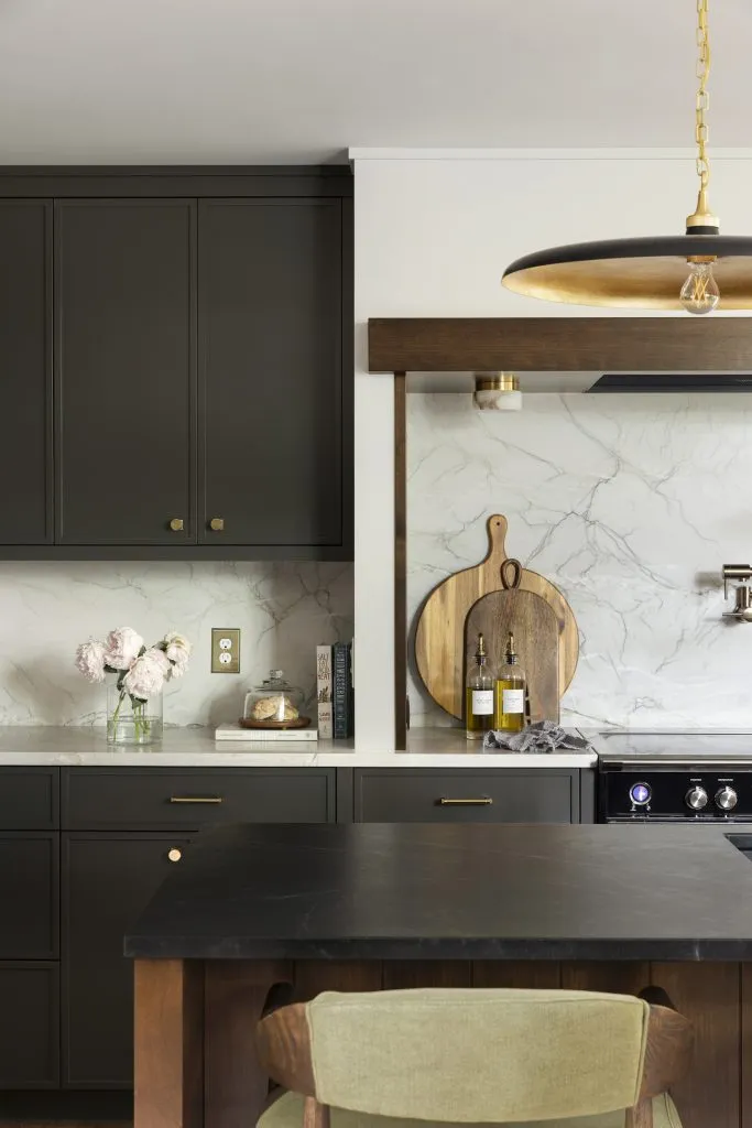 A modern kitchen with dark cabinets, marble backsplash, and a black countertop island—an inspiring space for those seeking A Guide to Cabinet Hardware Placement. Wooden accents, pendant lighting, and tasteful décor enhance the stylish atmosphere.