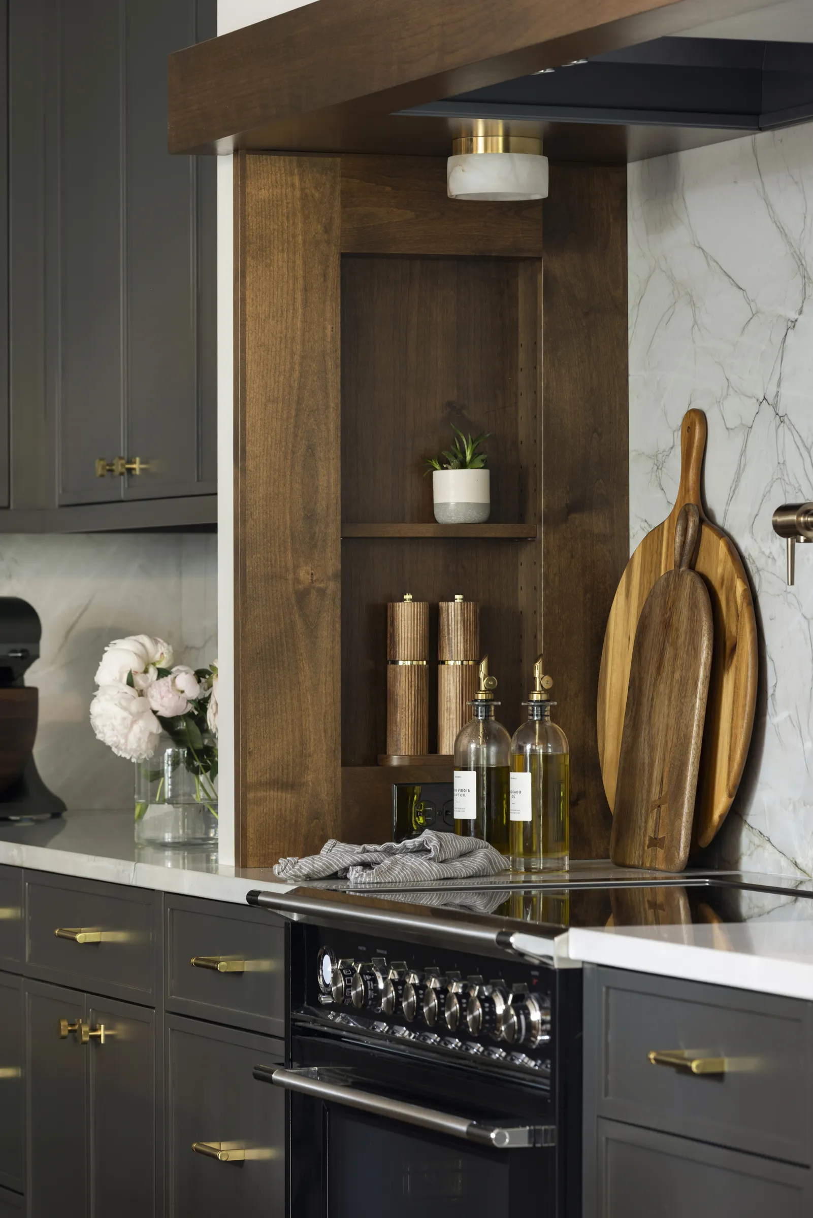 A modern kitchen with dark cabinets, gold hardware placed with precision inspired by A Guide to Cabinet Hardware Placement, white marble backsplash, and a black stove. Wood shelves display a potted plant and cooking essentials near wooden cutting boards.