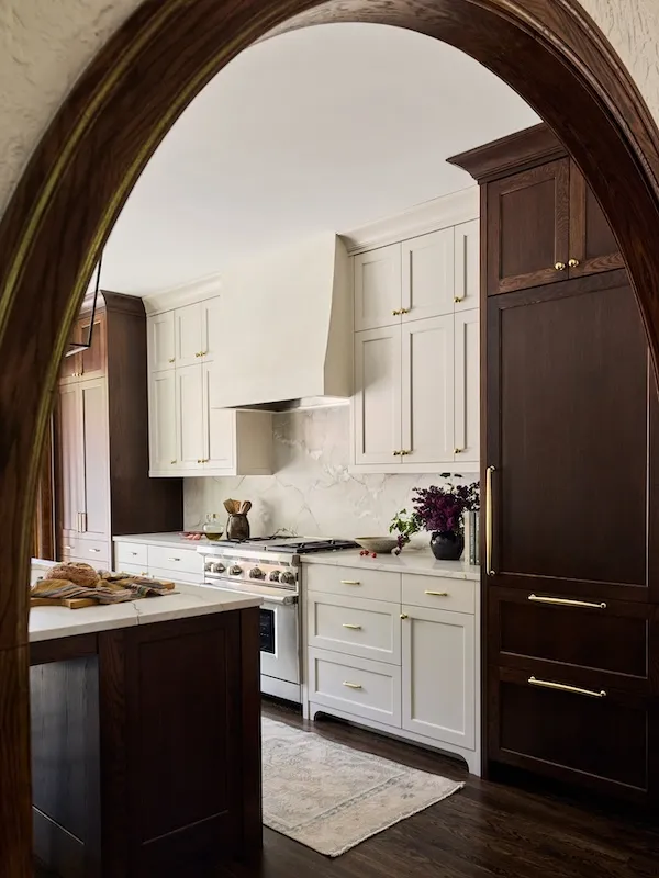A modern kitchen at Woolman Court is viewed through a wooden archway, featuring white cabinets, a marble backsplash, dark wood cabinetry, gold hardware, a range stove, a small rug, and a vase with dark purple flowers on the countertop.