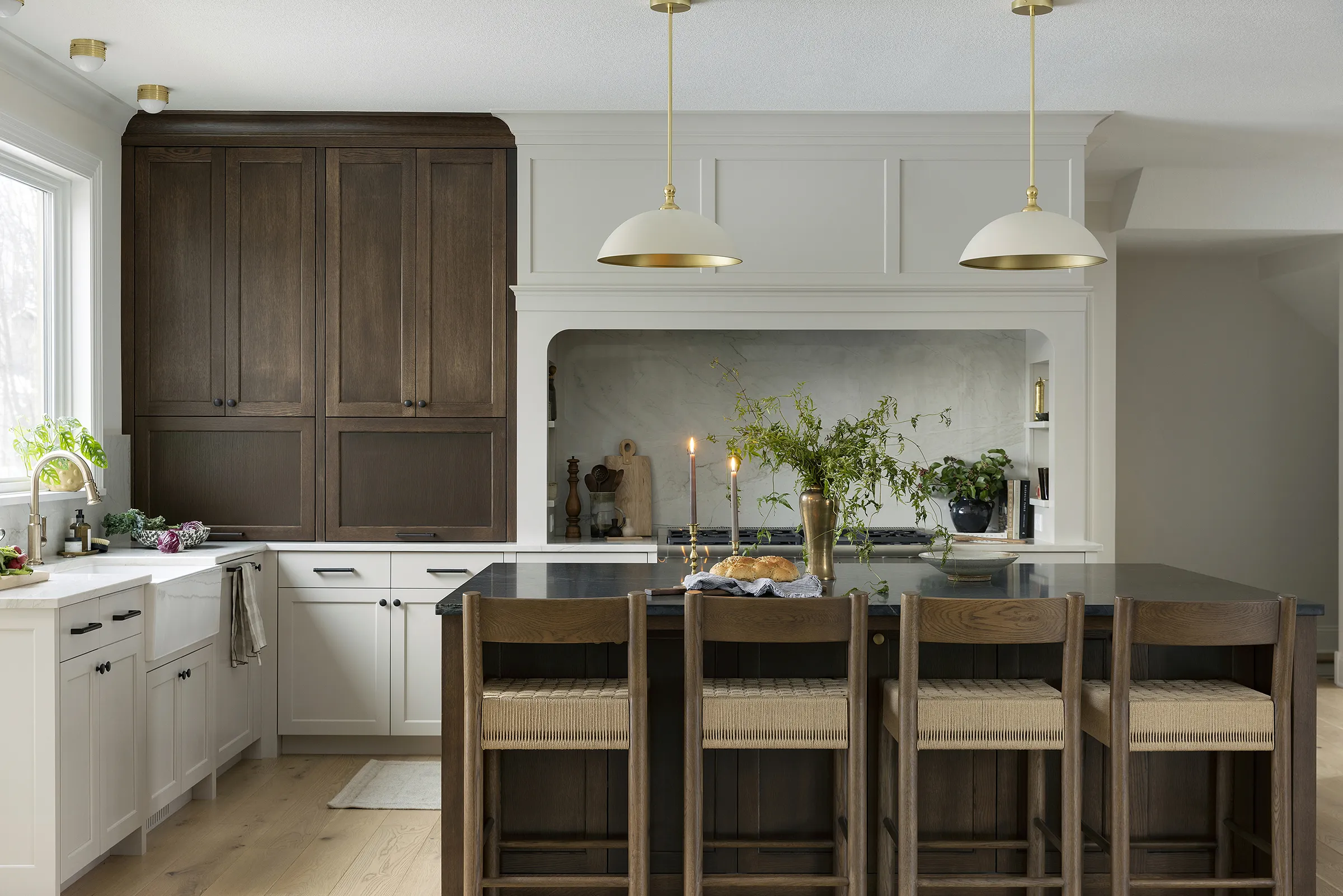 A modern kitchen with white cabinets, dark wood accents, and a large island with four woven-seat chairs exudes Bungalow Refined charm. Pendant lights hang above, while greenery and candles decorate the countertop as natural light streams in.