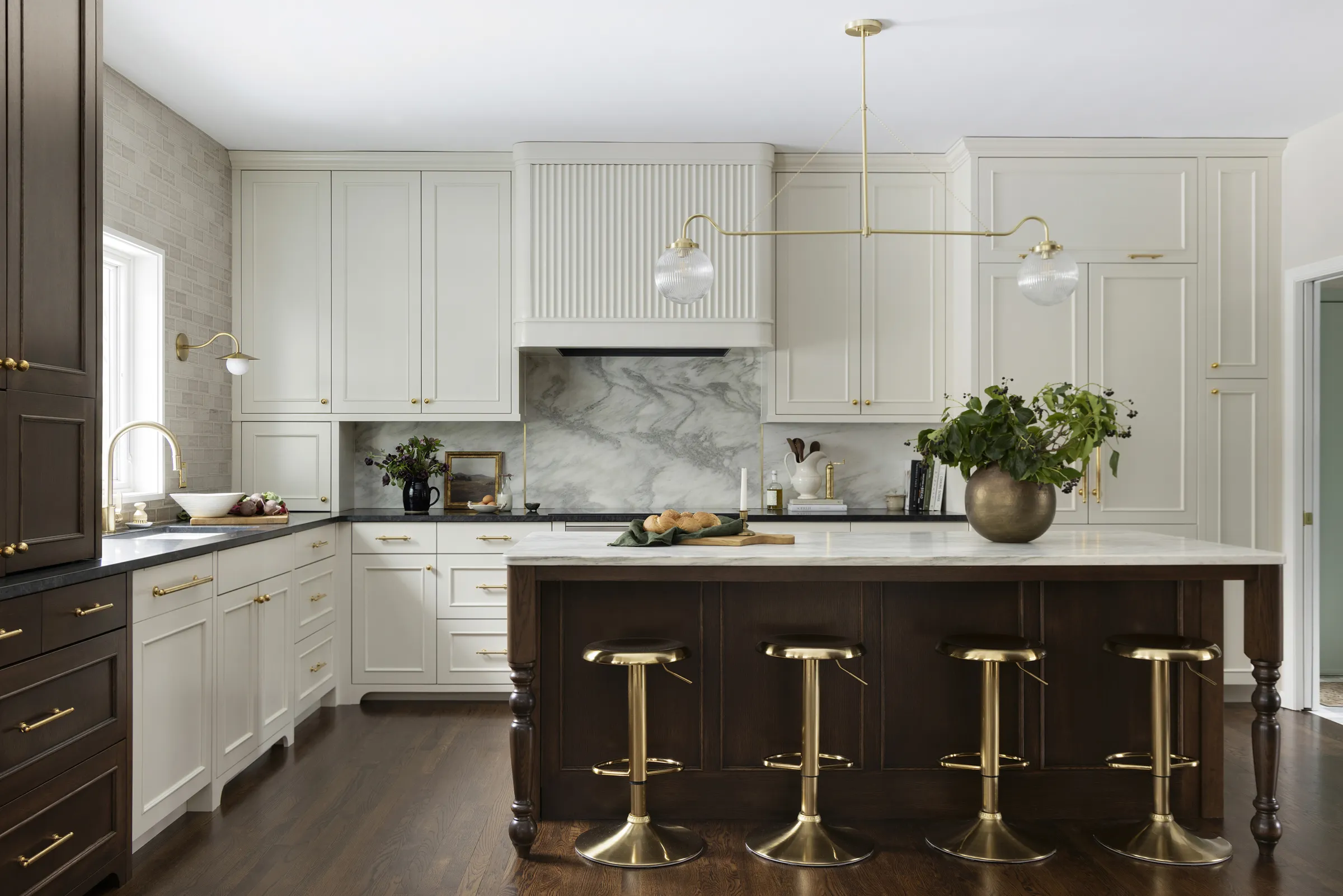 This kitchen renovation has creamy off white painted cabinetry that wraps the room, letting proportion and detail do the heavy lifting. A reeded hood feels architectural rather than decorative, paired with a full-height marble backsplash that reads as one continuous, honest (see above) surface. The dark-stained island grounds the space like a piece of furniture; turned legs, warm wood, no apologies, while brass brings just enough glow to this brand new kitchen.