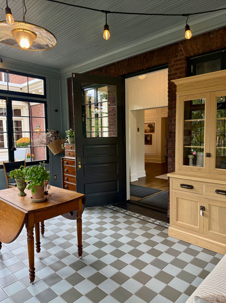 A bright sunroom with checkered tile floor, wood furniture, potted plants on a table, glass-front cabinet, string lights, and a Dutch door open to a hallway with art and hardwood floors—an inviting space in A House That Grows With You.