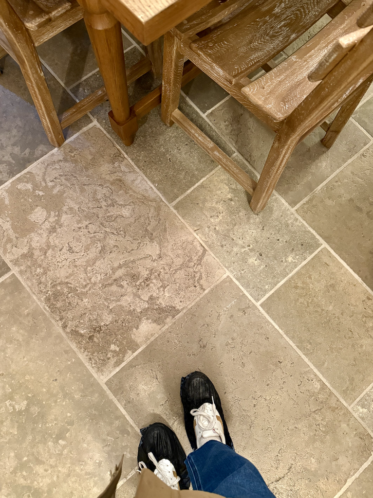 A pair of feet in black and white sneakers stand on beige tiled flooring next to wooden chairs and a table, capturing a quiet moment in A House That Grows With You.