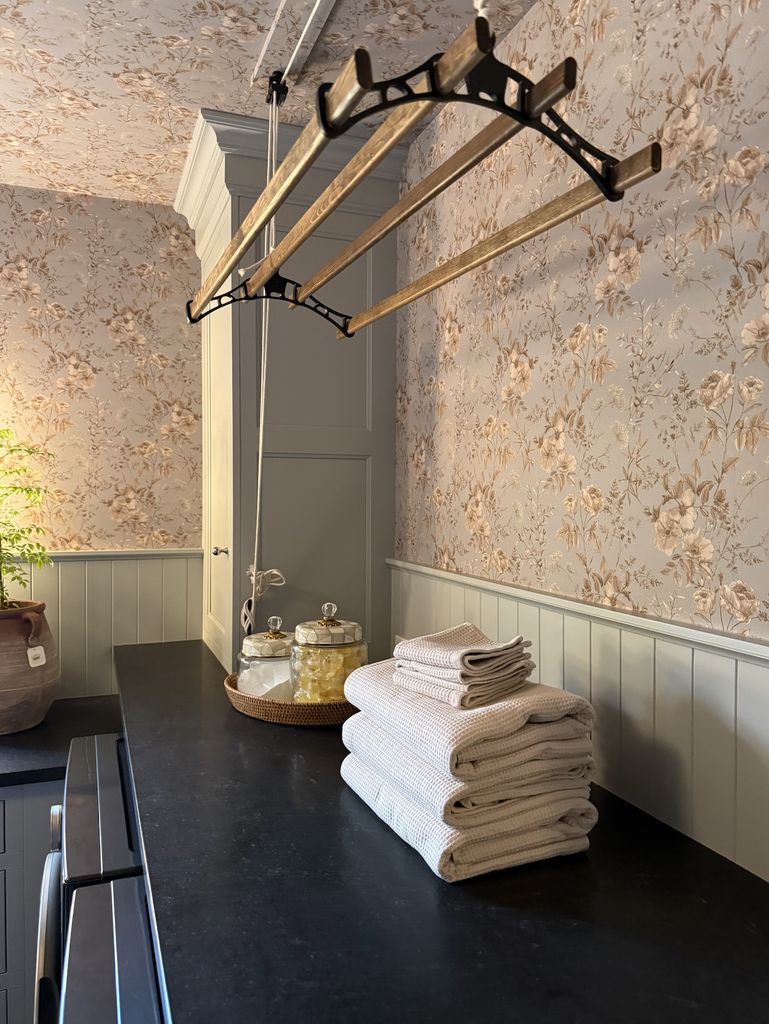 A vintage-style laundry room with floral wallpaper, a black countertop, folded towels, glass jars, and a wooden drying rack suspended from the ceiling. Soft natural light brightens the cozy space—an inviting blend of practicality and charm in A House That Grows With You.