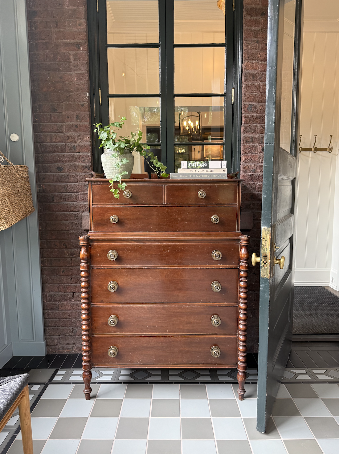A wooden dresser with brass handles stands against a brick wall in A House That Grows With You. A green potted plant and books sit on top, adding cozy character. Behind it, a glass window reflects lights, while an open door to the right and tiled floors complete the inviting scene.