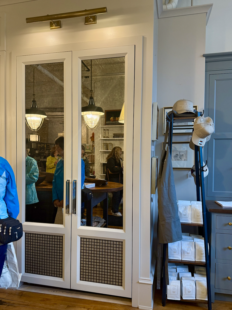 A pair of glass double doors reflect people inside a cozy shop with warm lighting, evoking the feeling of A House That Grows With You. To the right, a rack displays hats, aprons, and art prints above shelves with packaged goods.