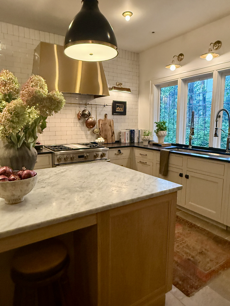 A House That Grows With You: A cozy kitchen with marble countertops, a large pendant light, gold range hood, white cabinets, a vase with hydrangeas, and a window overlooking trees. A stove, books, and plants are on the counter—creating a warm space designed to adapt over time as your needs change.