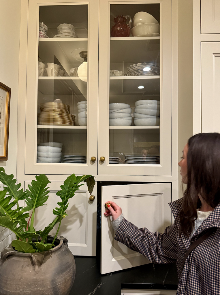A woman opens a lower cabinet in a kitchen. Above her, glass-front cabinets display neatly stacked white dishes and bowls. A large potted plant sits on the dark countertop, infusing life into A House That Grows With You.