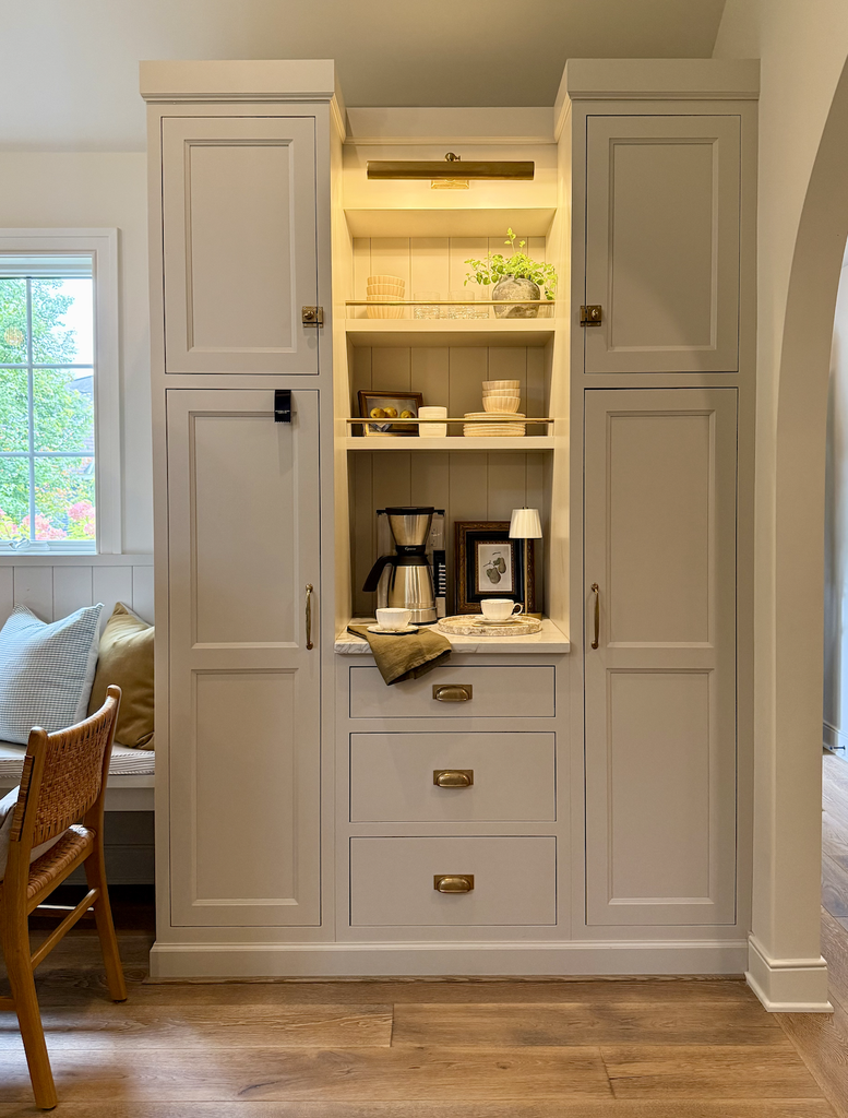 Built-in beige cabinet with shelves holding a coffee maker, cups, books, and decor. Drawers below, overhead light, and cabinet doors frame the display—a thoughtful design choice for A House That Grows With You. A chair and window are partially visible on the left.