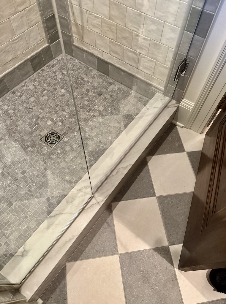 A House That Grows With You: A glass shower with gray and white tile, a square drain on the floor, and a checkered bathroom floor outside the shower. Part of an open wooden door is visible—designed for style and adaptability as your needs change over time.