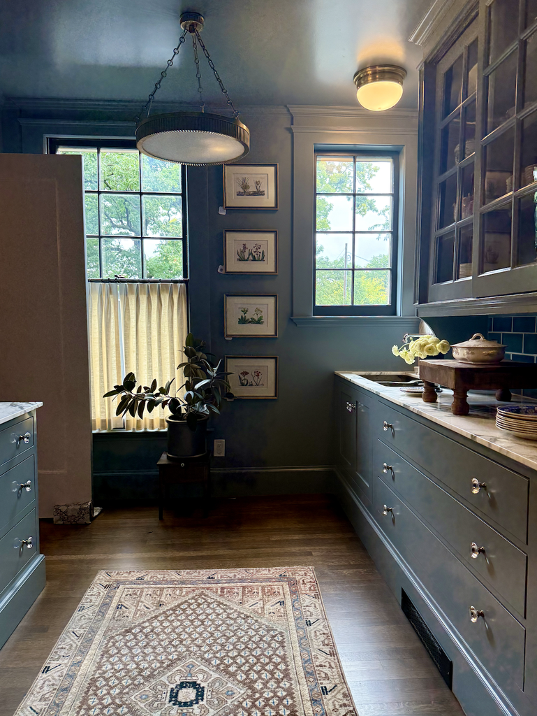 A cozy kitchen with blue cabinets, wooden countertops, plants, framed art on the wall, a window with a half curtain, and a patterned rug on the floor. Warm lighting creates an inviting atmosphere—perfect for A House That Grows With You.